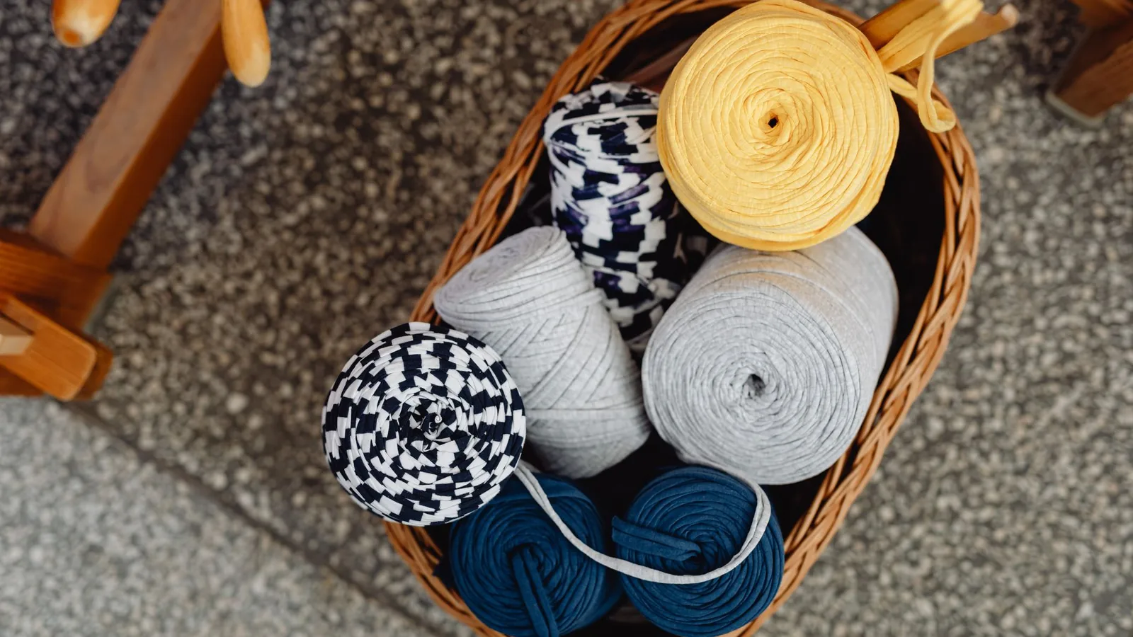 A wicker basket of yarn rolls in blue, grey and yellow beside a wooden chair leg.