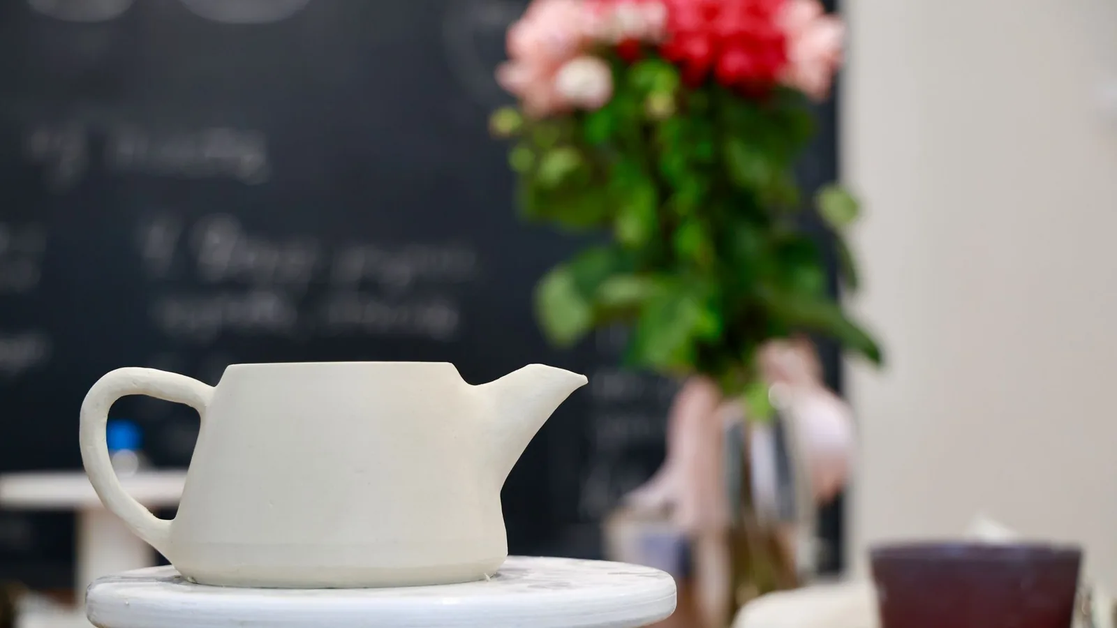 A plain white teapot on a table, with a vase of pink and red flowers behind it.