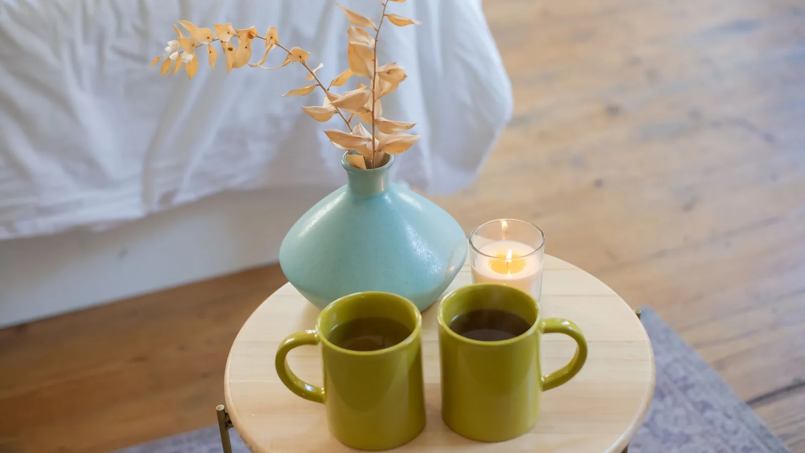 Two green mugs of tea on a small wooden table beside a vase and a lit candle