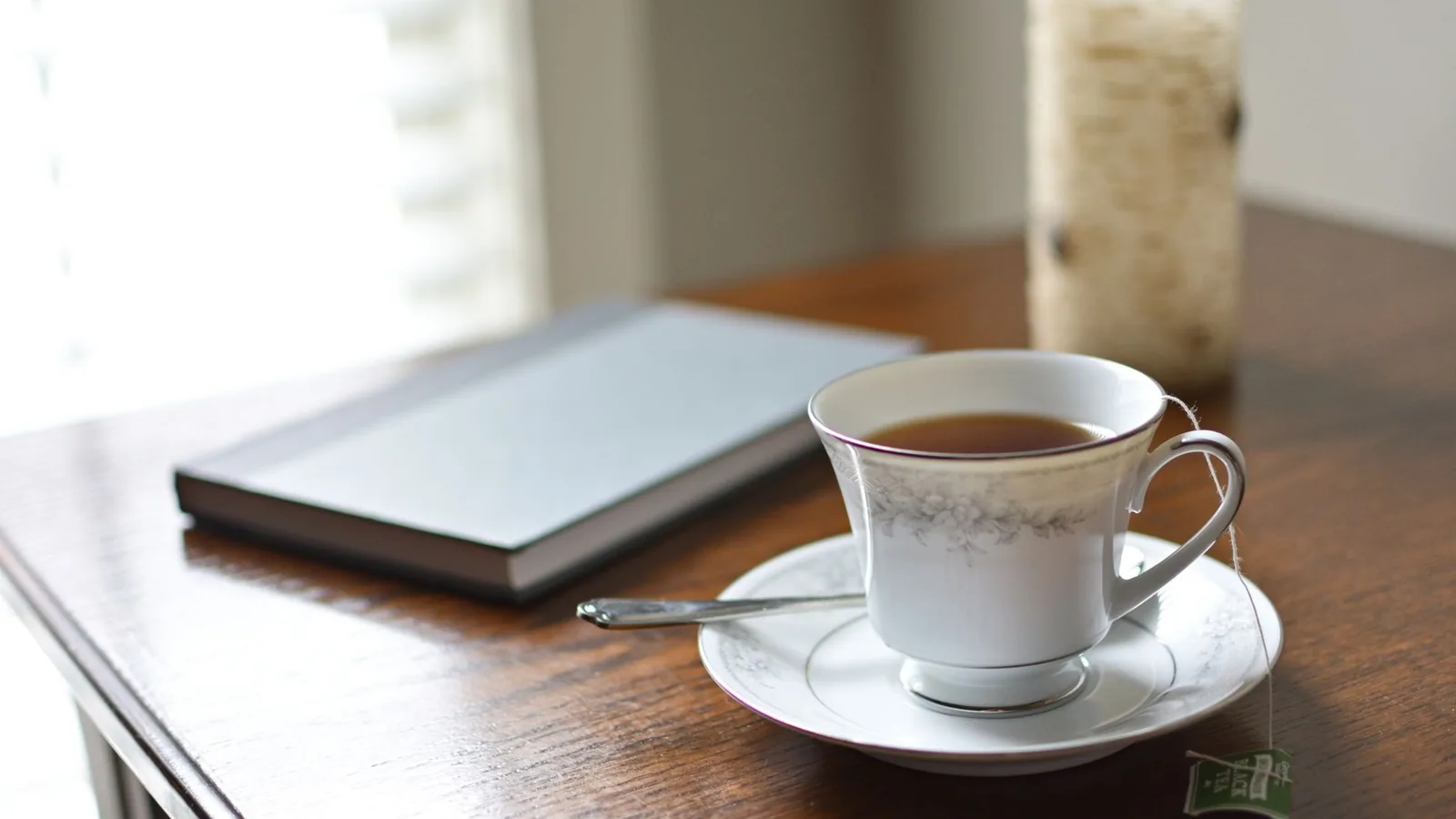 A china cup of tea on a saucer beside a closed notebook on a wooden table near a bright window.