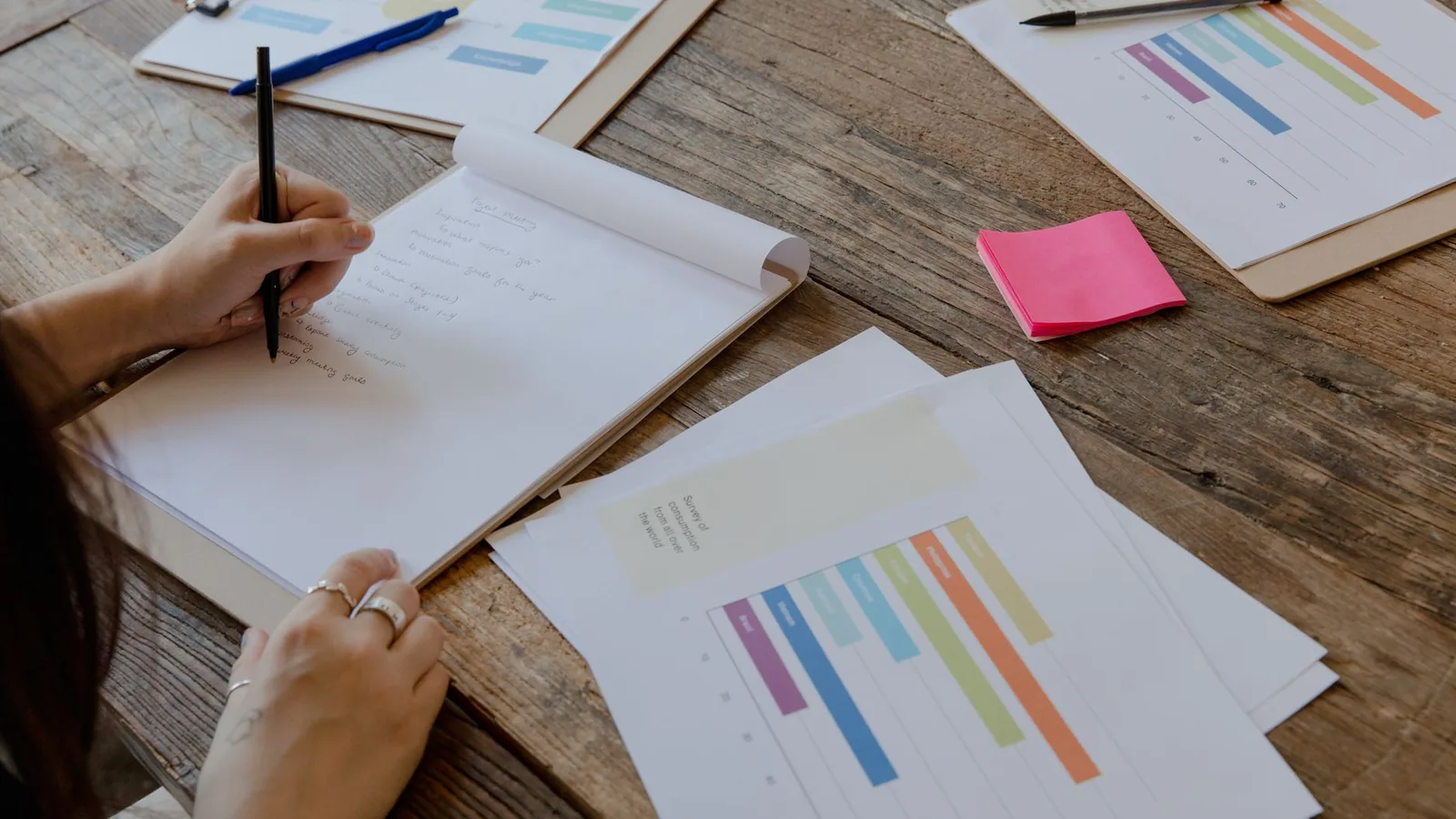 Hands writing notes on a pad beside printed bar charts and a pink sticky note on a wooden table.
